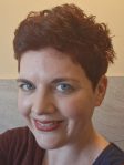 Headshot of a woman in her 30s with short curly red-brown hair and blue eyes smiling at the camera.